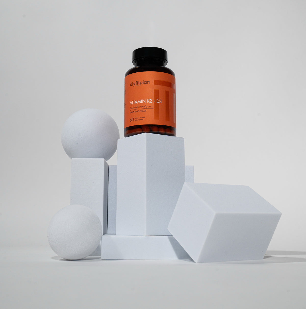 Supplement bottle labeled 'Vitamin K2 + D3' on geometric white blocks with a gray background