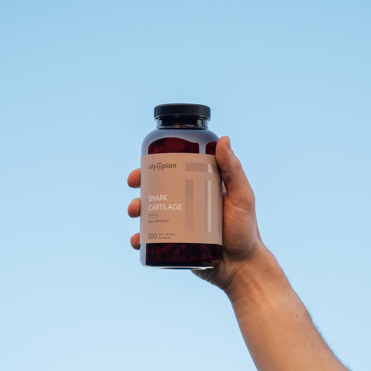 Hand holding a bottle of 'Olympian' shark cartilage supplement against a clear blue sky.