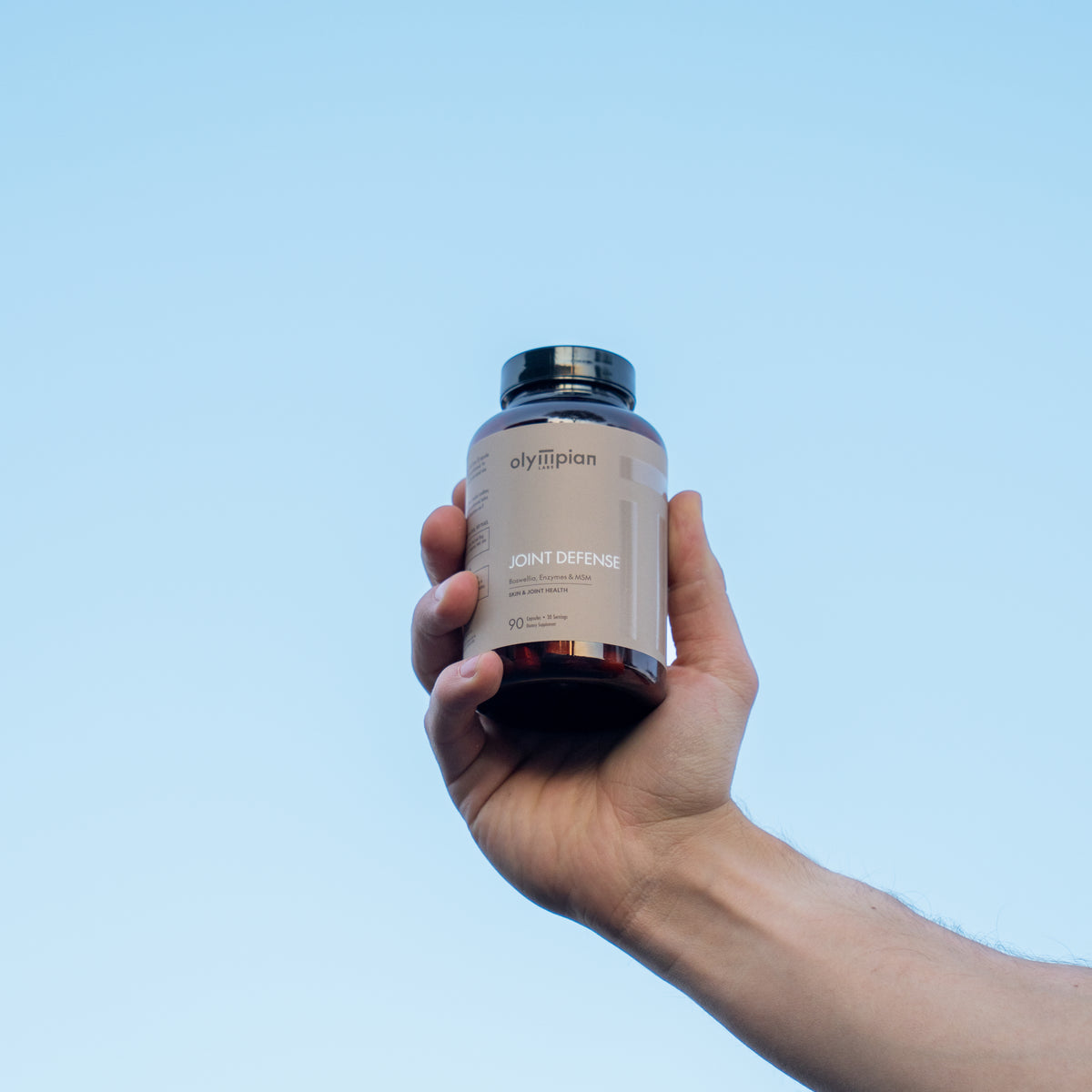 Hand holding a supplement bottle labeled 'Joint Defense' against a clear blue sky