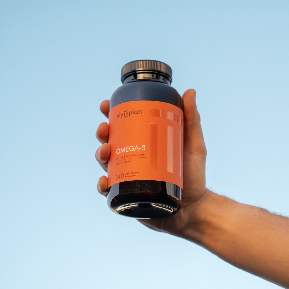Hand holding a bottle of Olympian Omega-3 supplement against a clear blue sky.